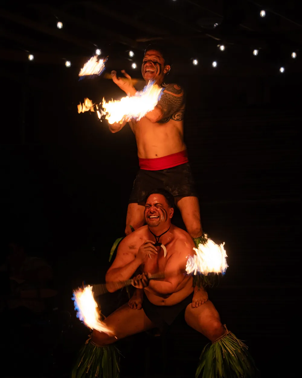 Two Polynesian fire performers laughing and smiling mid-act, one balanced on the other's shoulders (representative photograph of the comic, crowd-playing side of a fire show, not Chief's Luau)